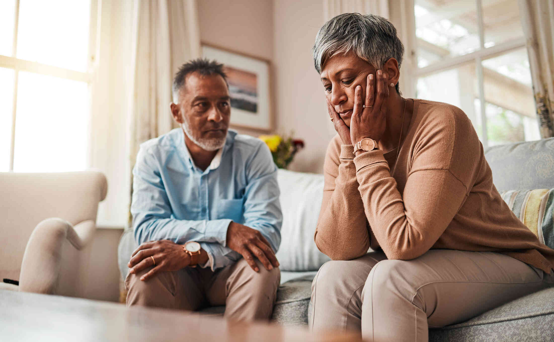 A middle aged wman sits hunched over on the couch with her head resting sadly in her hands as her male partner sits nearby with a worried expression.